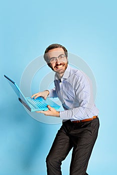 Cheerful Caucasian man, businessman in office dress code having fun, dancing with laptop over blue background. Positive
