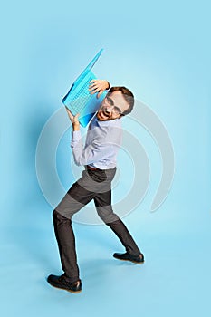 Cheerful Caucasian man, businessman in office dress code having fun, dancing with laptop over blue background. Positive