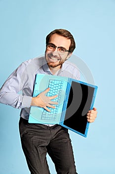 Cheerful Caucasian man, businessman in office dress code having fun, dancing with laptop over blue background. Positive