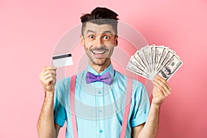 Cheerful caucasian man with moustache and bow-tie showing plastic credit card with money in dollars, smiling at camera