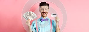 Cheerful caucasian man with moustache and bow-tie showing plastic credit card with money in dollars, smiling at camera
