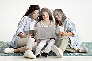 Cheerful caucasian teen girl, adult mother and old grandmother with gadgets sit on floor