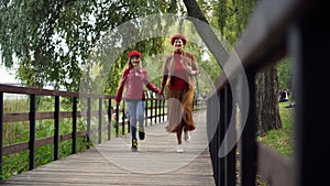 Cheerful Caucasian woman and girl jumping strolling holding hands having fun in autumn park. Wide shot portrait of