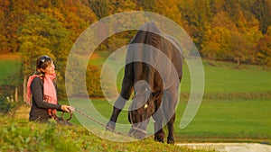 Cheerful Caucasian young woman watches her horse grazing on a calm morning.