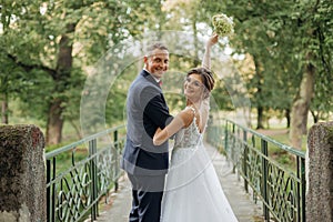 Cheerful, chancy young married couple of man and woman in glasses standing together on bridge in park, holding hands