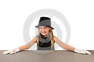 Cheerful charming little eight-year-old magician girl in hat and gloves is preparing to show tricks and presentations at gray tabl
