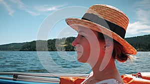 Cheerful charming woman in straw hat floating on boat, enjoying beautiful nature