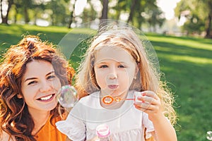 Cheerful child blowing soap bubbles in the park with woman looking at the camera