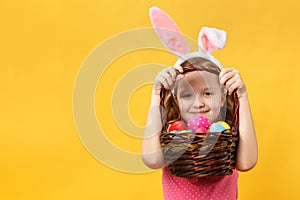 A cheerful child in the Easter ears of a bunny peeks out from under the handle of a basket. Little girls on a yellow background
