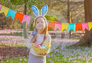 cheerful child girl with rabbit ears on her head and a basket with Easter eggs at party in garden