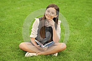 cheerful child in glasses reading book sitting on green grass