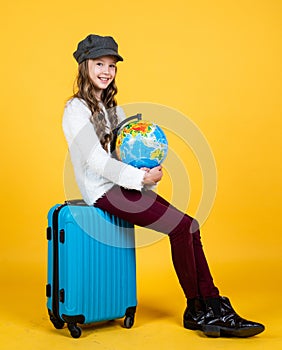 cheerful child in headwear with valise for travelling on yellow background, holiday