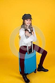 Cheerful child in headwear with valise for travelling on yellow background, vacation