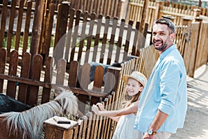 cheerful child and man smiling while standing near pony in zoo.