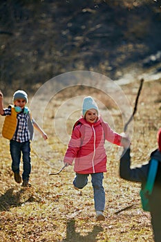 Cheerful child in pink jacket with wood stick leads playmates in lively run across sunlit field. Outdoor activities