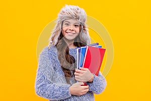 cheerful child in sweater and earflap hat with workbook on yellow background, education