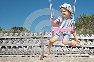 A cheerful child swings on the beach as he looks into the distance. A sunny summer day, blue skies and a happy childhood