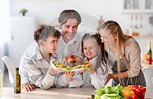 Cheerful children with parents cooking traditional dinner together, holding roast turkey for holiday meal at kitchen