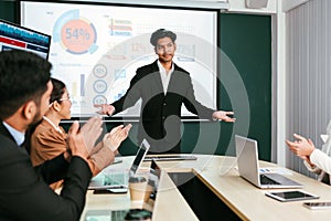 A cheerful and confident Asian businessman stands, presenting bar charts data from the projector screen to his office colleagues.