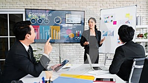 A cheerful and confident Asian businesswoman stands, present bar charts data from a whiteboard to her office colleagues.