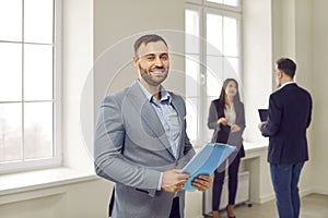 Cheerful confident businessman standing in office on background of colleagues