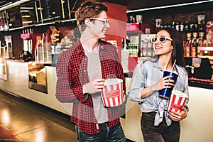 Cheerful couple in glasses are looking to each other and smiling. They are posing. Girl has a cup of coke and a basket
