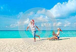 Cheerful couple in red Santa hats walking at tropical ocean sandy beach with sleds, decorated fir-tree and gift boxes. Happy New Y