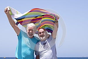 Cheerful couple of senior bearded brothers laughing at the beach holding a colorful towel in the wind - concept of freedom and