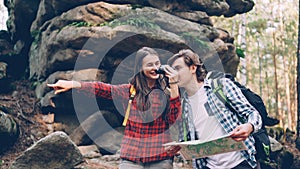 Cheerful couple is standing in forest, man is studying map, girl is looking through binoculars then showing interesting