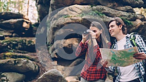 Cheerful couple is standing in forest, man is studying map, girl is looking through binoculars then showing interesting