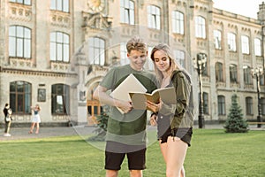 Cheerful couple of young people are standing on the lawn on the background of a college building with a laptop and noteboo,