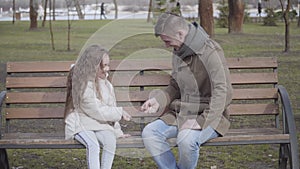 Cheerful curly-haired girl playing rock-paper-scissors game with father in city park. Happy young man sitting on bench