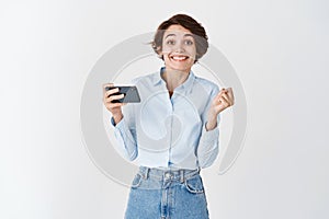 Cheerful cute girl winning on mobile phone, making money in smartphone app and smiling, standing happy on white