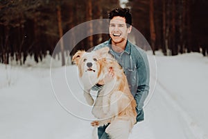 Cheerful cute laughing and smiling guy in jeans clothes with dog border collie red on his hands in snowy forest. concept