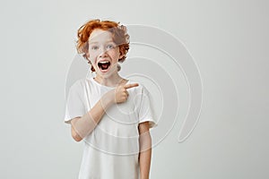 Cheerful cute little boy with curly ginger hair and freckles happy smiling and pointing aside with finger on white