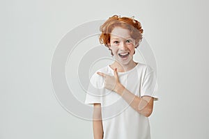 Cheerful cute little boy with curly ginger hair and freckles happy smiling and pointing aside with finger on white