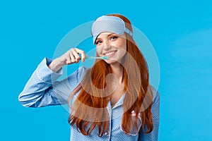 Cheerful cute redhead woman in nightwear, pyjama and sleep mask, brushing teeth holding toothbrush and smiling cheerful