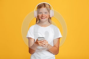 Cheerful cute teen blonde girl in headphones use phone and looks at camera