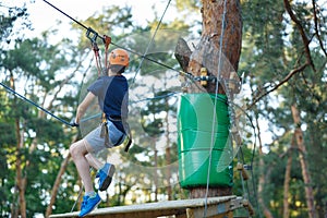 Cheerful cute young boy in blue t shirt and orange helmet in adventure rope park at sunny summer day. Active lifestyle, sport