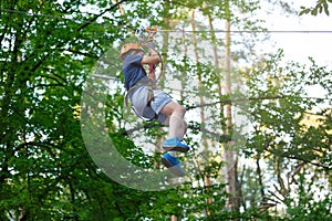 Cheerful cute young boy in blue t shirt and orange helmet in adventure rope park at sunny summer day. Active lifestyle, sport