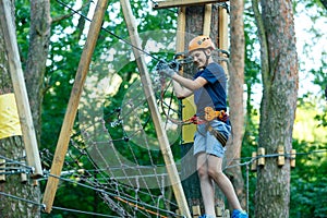 Cheerful cute young boy in blue t shirt and orange helmet in adventure rope park at sunny summer day. Active lifestyle, sport