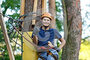 Cheerful cute young boy in blue t shirt and orange helmet in adventure rope park at sunny summer day. Active lifestyle, sport