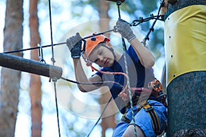 Cheerful cute young boy in blue t shirt and orange helmet in adventure rope park at sunny summer day. Active lifestyle, sport