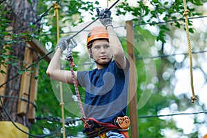 Cheerful cute young boy in blue t shirt and orange helmet in adventure rope park at sunny summer day. Active lifestyle, sport