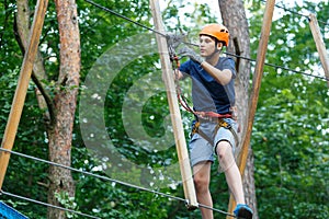 Cheerful cute young boy in blue t shirt and orange helmet in adventure rope park at sunny summer day. Active lifestyle, sport,