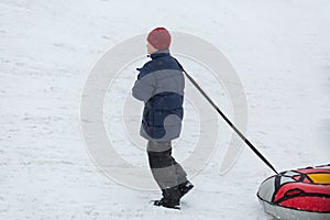 Cheerful cute young boy in orange hat red scarf and blue jacket holds tube on snow, has fun, smiles. Teenager on sledding