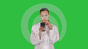 Cheerful cute young woman doctor typing text on phone while walking on a Green Screen, Chroma Key.