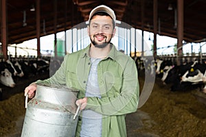 Cheerful dairy farm owner carrying can of milk in cowshed