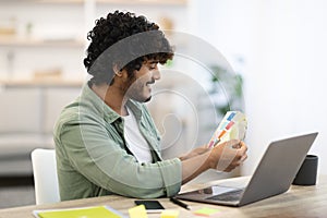 Cheerful dark-skinned guy sitting at workdesk in front of laptop