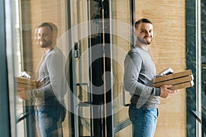 Cheerful delivery man holding boxes pizza and contactless payment POS terminal, standing in entrance hall of apartment